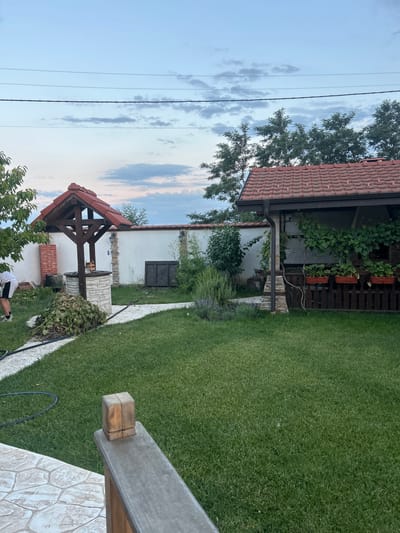 In Войводино, Bulgaria, a residential backyard is captured with a well, a covered structure, and lush green grass. A wooden well structure with a red tiled roof stands prominently, surrounded by a stone base and a pile of trimmings to its left. Adjacent to the well is a white wall with two outdoor lights. To the right, a wooden structure with a red tiled roof features a fence with potted plants and climbing vines. The yard is meticulously maintained with a well-mown lawn. A person in black and white shorts is visible in the lower left corner, partially obscured by a tree, appearing to be working with a hose on the ground. The sky above is a soft blue with scattered clouds.