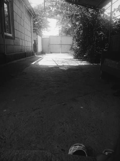 The black and white photograph captures an outdoor scene, likely a courtyard or driveway, in Taraz, Kazakhstan. Sunlight streams in from the background, creating strong shadows on the ground and illuminating a portion of the pathway leading towards a large metal gate. To the left, a wall constructed of concrete blocks forms part of a building, with a window visible. Lush green foliage encroaches from the right side of the frame, partially obscuring another structure. In the foreground, a roll of what appears to be tape with the "NIKE" logo printed repeatedly is visible, along with a glimpse of a person's feet wearing slides also bearing the Nike logo. The strong contrast between light and shadow suggests it is daytime, possibly late morning or early afternoon, with clear weather. The overall mood is somewhat tranquil, with the play of light and shadow creating a dynamic visual effect.