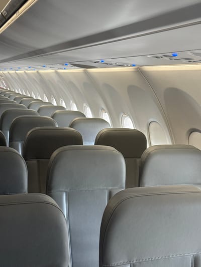The image depicts the interior cabin of a commercial aircraft, located in Tijuana, Mexico. Multiple rows of unoccupied passenger seats, upholstered in a uniform grey material with distinct horizontal stitch lines, are visible. The perspective is from behind several seats, looking forward and slightly towards the starboard side of the aircraft. The cabin features standard overhead luggage compartments and ceiling panels, some of which display illuminated blue indicator lights. A series of circular passenger windows is present along the right side of the fuselage, allowing natural light into the cabin. No individuals are present, and no specific actions or interactions are occurring. The environment is well-lit by both artificial cabin lighting and ambient light from the exterior.