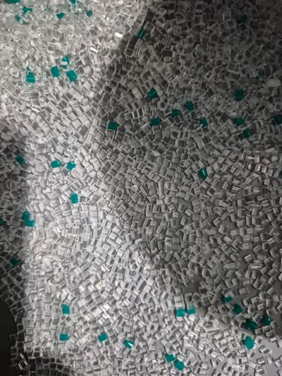 The image displays a close-up, overhead view of a large pile of translucent, square plastic pellets. Interspersed throughout the pile are a smaller number of opaque, teal-colored pellets. The lighting creates shadows and highlights on the pellets, suggesting they are in a container or on a surface. There are no people or discernable objects other than the plastic pellets. The scene is devoid of any environmental context that would place it in the city of Pabradė, Lithuania, or any other specific location.