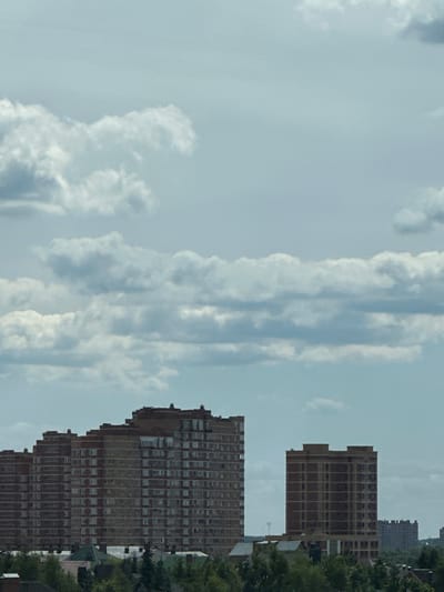 This media file depicts an outdoor scene of apartment buildings against a cloudy sky. The image is oriented vertically, with the sky occupying the majority of the frame. Several tall, brick apartment buildings are visible on the right side of the image. These buildings feature numerous windows arranged in a grid pattern. In the foreground, a large, multi-story apartment building dominates the lower right portion of the frame. Behind it and to its right, another similar building is partially visible. Further in the background, another, taller building with a darker facade can be seen, partially obscured by trees. The sky is filled with a mix of light and dark clouds, suggesting an overcast or partly cloudy day. The lighting appears diffused, consistent with daytime, but the exact time of day is not precisely determinable. There are no people or specific activities depicted in the image. The setting appears to be an urban or suburban residential area, likely in Znamya Oktyabrya, Russia, given the context provided. No text is visible in the image.