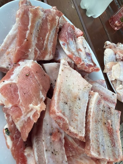 The image shows a close-up, overhead view of raw pork ribs and chicken wings on white plates, suggesting food preparation for a meal. The ribs are seasoned with black pepper and appear to be arranged in a layered fashion. The chicken wings are also visible on a separate plate. The background reveals a wooden surface, possibly a table, and a glimpse of a glass containing a pink liquid with some herbs or fruit. A white gloved hand is partially visible in the upper right corner, indicating someone is handling the food. The lighting is bright, highlighting the texture and marbling of the raw meat. There is no indication of the specific location being in Mordarka, Poland.