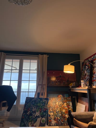 The image shows a room with a large window overlooking the Mediterranean Sea. The room is an artist's studio, filled with paintings, an easel, and art supplies. The room has a light blue wall and tan curtains framing the windows. The floor is wooden. Two large paintings, abstract in style, are positioned against the wall on the right. There is a wooden table in front of them, and on it are art supplies, a glass of water, and some cans.  A floor lamp with a white shade sits on the table, illuminating the area. A person's feet are visible sitting in a large, grey armchair, facing the window. One foot is on the floor and the other is resting on the seat of the armchair. The coordinates (latitude: 43.763, longitude: 7.464) correspond to the location of Roquebrune-Cap-Martin, France. This is the location of the artist's studio.