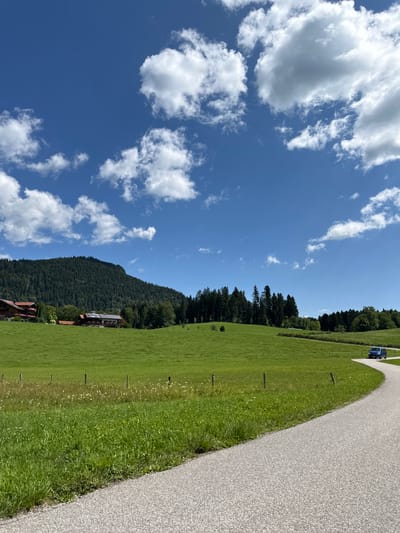 In Bischofswiesen, Germany, a paved road curves through a bright green, rolling meadow under a vibrant blue sky dotted with fluffy white clouds. The meadow is lined with sparse wooden posts and wire fencing. In the background, a dense forest covers a large, rounded mountain. To the left of the mountain, a few buildings with reddish-brown roofs are nestled amongst the trees and fields. A blue van is visible driving on the road in the distance, to the right of the frame. The overall scene depicts a tranquil, rural landscape.