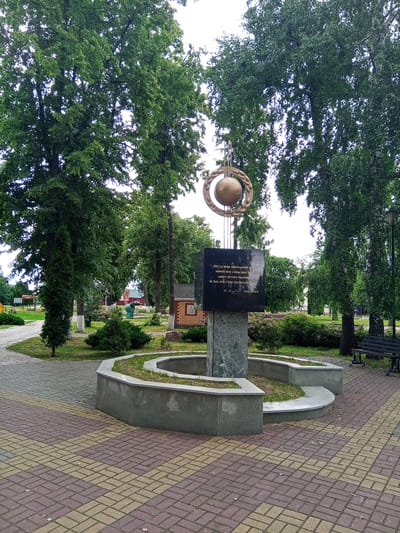 This media file is a photograph of a monument in a park. The monument, which appears to be made of granite and dark stone, features a circular metal design with a globe at its center. Inscriptions in Russian are visible on the dark stone portion. The monument is surrounded by a low, curved stone planter with grass. The park setting includes well-maintained trees, manicured bushes, and a paved walkway made of brick in a herringbone pattern. A park bench is visible to the right of the monument. In the background, there are more trees, a building with a red roof, and a playground structure. The sky is overcast, suggesting it is daytime with cloudy weather. The overall scene conveys a sense of peace and remembrance, likely in a public space dedicated to a historical figure or event. The text on the monument appears to be a quote.