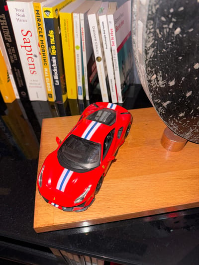 A top-down shot showcases a red toy car with a blue and white stripe, sitting on a wooden surface near a black countertop. The car is positioned next to a stack of books, including titles such as "Sapiens," "The Miracle Morning," "La Blockchain pour les nuls," and works by Stephen Hawking and Sigmund Freud. A patterned object partially visible in the upper left corner adds context to the scene.