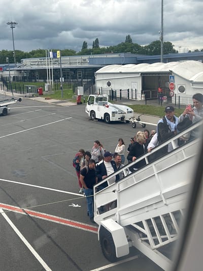 The image depicts an outdoor scene, likely at an airport or a large event venue in Tillé, France, under an overcast sky. In the foreground, numerous people, mostly adults, are congregated around and on the steps of a white, wheeled passenger stair unit. They appear to be in the process of boarding or disembarking, or waiting to do so. The individuals are dressed casually.

The paved ground features white lane markings, a prominent orange-red stripe, and a distinctive white stylized figure resembling a person. In the midground, several white utility vehicles, typical of airport ground support, are visible. One such vehicle has partially visible text, possibly "IID," on its side.

The background consists of large, white tent-like structures or temporary buildings, some fenced off. Beyond these, more permanent buildings with large glass sections and a dense line of green trees are visible beneath the heavy, grey clouds.

Notable details include several road signs near the white tents: a red octagonal "STOP" sign, a blue square information sign with an upward arrow, and a red circular prohibitory sign. Further back, two flags fly from poles: one is the blue and yellow-starred flag of the European Union, and the other is a yellow flag with a black number "2." The cloudy conditions indicate daytime.