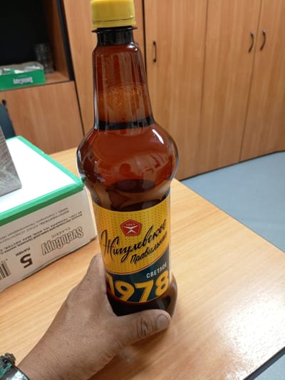 A hand is holding a brown plastic bottle with a yellow cap and a yellow and blue label. The label has text in Russian, including "Хипулевское" and "1978". A portion of a cardboard box with the word "SvetoCopy" printed on it is visible in the lower left of the image. The background consists of a wooden desk, a wooden cabinet with two doors and handles, and a portion of a wall. The scene appears to be indoors, likely in an office or storage area.