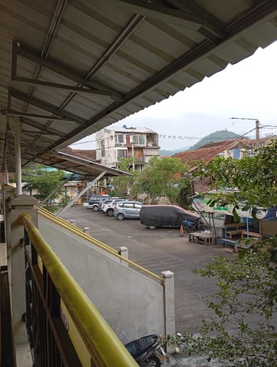 The image displays an urban outdoor scene in Pamekasan, Indonesia, under an overcast sky. The foreground is dominated by the underside of a corrugated metal roof structure, supported by metal beams, extending from the upper left. Below this, a light grey concrete balustrade with a yellow-painted top railing and a dark metal lattice railing is visible. A dark scooter is partially visible at the base of the balustrade. The midground features a paved area serving as a parking lot, containing a dark blue SUV, a white car, a silver hatchback, and a dark-colored vehicle completely covered by a black tarpaulin. To the right, two wooden tables and two wooden benches are situated near an informal vendor stall constructed with blue and green tarpaulins. Green foliage from trees and bushes is scattered throughout. In the background, multiple buildings with reddish-brown tiled roofs are present, along with a prominent, multi-story, weathered building featuring various windows and balconies. Utility poles, power lines, and a distant green hill range are visible on the horizon. No individuals are present, and no actions or interactions are depicted.