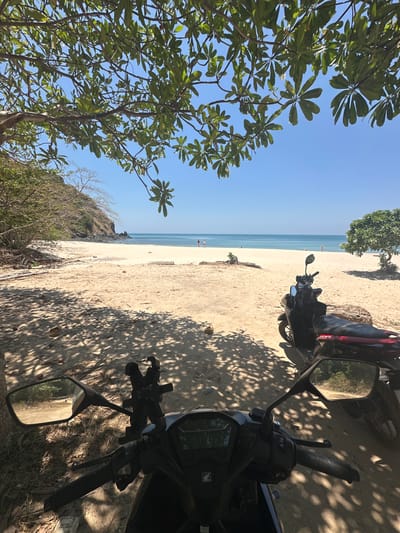The image displays a sunny beach scene at Ao Khlong Jak, Thailand. The foreground is dominated by the handlebars and dashboard of a dark-colored Honda scooter, equipped with a black phone mount and visible rearview mirrors. The ground directly beneath is light brown sand, covered in distinct dappled shadows cast by overhead tree foliage. Partially visible behind the first scooter is the rear section of a second dark-colored scooter, featuring a red seat, also parked on the sand.
The midground reveals a broad expanse of light-colored sandy beach extending towards the ocean. Scattered pieces of driftwood or fallen logs are present on the sand. On the right side of the beach, a small, full-leafed tree adorned with white flowers stands near the sand.
The background shows the calm, clear blue ocean meeting the sandy shore under a bright, cloudless blue sky. Multiple distant human figures are discernible on the beach near the water's edge; two individuals are positioned together on the left-center, and another figure stands further right. To the far left, a vegetated hillside, dense with trees and shrubs, rises directly from the beach line. The entire scene is illuminated by bright natural sunlight, indicating a clear day.