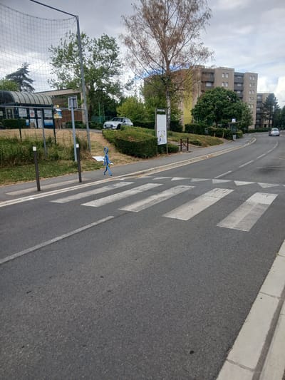 The image captures a street scene in Charleville-Mézières, France. The perspective is from a high angle, looking down onto a pedestrian crossing with white stripes on an asphalt road. The road curves gently to the left.
In the background, several multi-story residential buildings stand, with visible windows and balconies. Lush green trees and hedges line the street and the grassy area next to the sidewalk. A few cars are parked or driving on the road, including a white Fiat 500.
On the sidewalk, a blue, stick-figure-like art installation is positioned near the pedestrian crossing. A black metal fence with netting, possibly part of a sports court, is visible in the lower left corner. There are also lampposts and what appear to be information boards with some text that is not clearly legible due to the image quality and angle.
The sky is overcast with some bright patches, suggesting a partly cloudy day. The time of day appears to be daytime, likely afternoon, given the lighting. The scene is calm and urban, with a mix of residential buildings, greenery, and public infrastructure. No people are clearly visible.