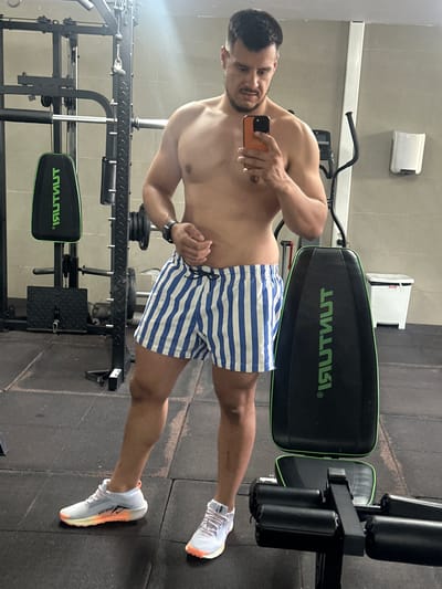 A man takes a selfie in a gym. He is shirtless and wearing blue and white striped shorts and white athletic shoes with orange accents. He is holding an orange-cased smartphone in his right hand. He has short dark hair and a beard and appears to be smiling or grimacing at the camera.

The man is standing in front of gym equipment, including a black weight bench with green trim and a logo that reads "ISULUTNUT" in a mirrored fashion. To his left, another piece of gym equipment, a rack with weights, is visible. The floor is black rubber matting. The walls are tiled in a light color.

The image is a vertical shot and appears to be taken indoors, likely in a gym in Roquetas de Mar, Spain. There are no explicit cues for time of day or weather. The overall mood is casual and focused on the man's physique.