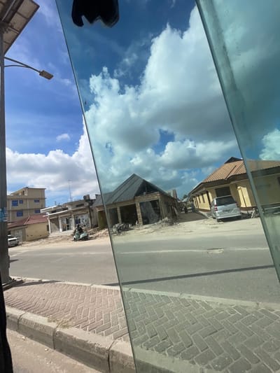 The image captures a complex, vertically oriented view from inside a vehicle, showcasing an outdoor street scene alongside significant reflections of the sky. The photograph is bisected horizontally by a prominent, distinct line, likely the edge of a car window or a strong reflection boundary.

The left side of the image is dominated by the sky. In the upper-left section, there is a clear blue sky filled with large, white, fluffy cumulus clouds. This area appears to be seen through a slightly tinted or smudged glass surface, as faint streaks and a greenish-blue hue are visible. The lower-left section also features a similar blue sky with white clouds. In the bottom-left corner of this section, a dark, amorphous shape is reflected, which could be part of the vehicle's interior, a person's head, or another object inside the car. Emerging from the very bottom edge of this section and curving upwards is a black streetlight pole with its lamp fixture clearly visible against the sky.

The right side of the image presents an outdoor street scene. A grey asphalt road runs vertically through the middle of these right-hand panels, suggesting it's a two-lane street. To the right of the road, there is a raised concrete curb and a sidewalk paved with grey brick pavers laid in a distinct herringbone pattern.

Along the right side of the road, various buildings line the street. In the upper-right portion, there is a light-colored, possibly yellowish or beige, building with a reddish-brown tiled roof. A white van or SUV is parked directly in front of this building, facing away from the viewer. Below this, closer to the center of the image, another structure is visible under construction, characterized by its exposed concrete or brick framework and a dark, likely metal, roof. Evidence of construction materials or debris can be seen around its base. Further down the street in the lower-right section, more buildings are present, including one multi-story building featuring several prominent blue-tinted windows. Patches of blue sky with white clouds are also visible above and between these buildings.

The scene appears to be set during daytime, under bright, clear skies with scattered clouds. The mix of finished and unfinished buildings suggests an urban or semi-urban environment, possibly a developing area or suburb. The overall perspective strongly indicates the photo was taken from inside a moving or stationary vehicle, looking out a side window. No discernible text is visible anywhere in the image.