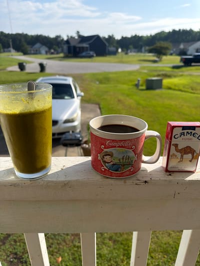 The image depicts a casual outdoor scene, likely on a porch railing, with a view of a suburban neighborhood in Elizabeth City, United States. The time of day appears to be late morning or early afternoon, given the bright sunlight casting shadows. The weather seems to be clear and sunny.
In the foreground, resting on a white, weathered wooden railing, are three objects. Closest to the viewer is a tall, clear glass filled with a greenish-yellow liquid, possibly a smoothie or juice, with some residue on the inside of the glass. Next to it is a red and white ceramic mug filled with dark liquid, likely coffee or tea. The mug has a design that includes a cartoonish depiction of a cowboy and possibly farm animals. To the right of the mug is a red and white box of Camel Filters cigarettes, featuring the brand's iconic camel logo.
In the background, out of focus, is a typical suburban street scene. There are houses with lawns, trees, and a white vehicle parked in a driveway. The overall impression is one of a quiet, everyday moment, perhaps someone enjoying a morning beverage and a cigarette while taking in the view. There is no visible emotion displayed, and the scene is static.
Visible text includes "CAMEL FILTERS SINCE 1913 TURKISH & DOMESTIC BLEND" on the cigarette pack, and "Campbell's" written on the mug.