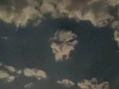 The image is a close-up photograph of a cloudy sky, likely taken during the daytime. The clouds are a mix of light, fluffy white and darker, more ominous gray tones, suggesting a partly cloudy or overcast sky. In the center of the frame, a formation of clouds appears to resemble a human face, with distinct features like eyes, a nose, and a mouth. This cloud formation is highlighted against the darker background of the sky. The overall impression is of a pareidolia effect, where the viewer's mind interprets random shapes as familiar objects or faces. The image does not depict any specific urban landscape or identifiable landmarks that would definitively place it in Қарғалы, Kazakhstan, or any other city. It is a natural phenomenon captured photographically.