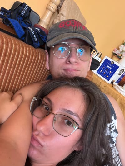 The image shows a close-up of two people, likely a couple, lying down together. One person is wearing a gray baseball cap with red lettering and glasses. The other person is wearing glasses and has their arm around the first person. They appear to be on a couch with an orange, striped texture. In the background, there are a few objects, including what looks like a package of coffee pods and a wooden carved object. The overall setting suggests an indoor environment. The image does not provide any information about the city of Golemo Buchino, Bulgaria, or any specific forensic details.