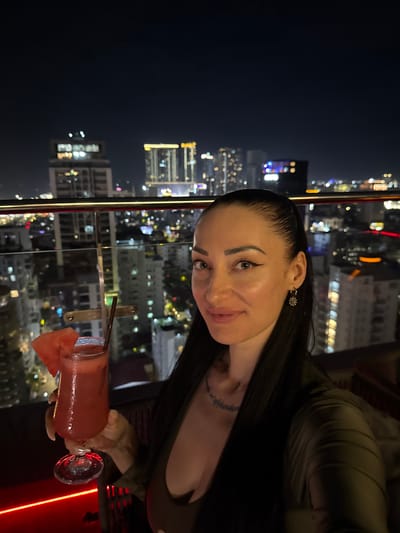 A woman with long, dark hair is holding a red cocktail with a slice of watermelon on the rim in a glass. She is wearing a dark top and has delicate earrings. A tattoo is visible on her neck. The scene appears to be a rooftop bar at night, with a blurred cityscape in the background featuring illuminated buildings. The time of day is night, and the weather is likely clear as the city lights are visible. The woman seems to be enjoying herself, with a slight smile on her face. The location is Khan Boeng Keng Kang, Cambodia. There is no visible text in the image.