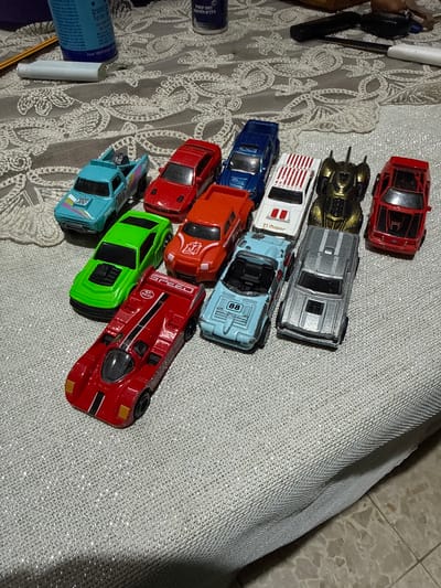 This image features a collection of toy cars arranged on a textured, light-colored fabric surface, possibly a tablecloth or blanket, with a hint of a tiled floor in the upper right corner. The fabric has a shimmering, reflective quality due to embedded threads or glitter, and a delicate white lace pattern is visible beneath the toys in some areas.
The toy cars are diverse in color and design. At the top, a red car with black accents is visible. Below it, a gold and black Batmobile-like vehicle is positioned. Next are a white car with red stripes and a blue car. Following these are an orange car, a bright green car, and a light blue car. The bottom of the arrangement is dominated by a red race car with black and white racing stripes and the word "SPEED" printed on its side. A dark blue spray can with Hebrew text and a white cylindrical object, possibly a cap or another spray can, are partially visible in the bottom left corner.
The lighting appears to be artificial, casting subtle shadows. There are no people present, and no specific activity is taking place other than the arrangement of the toys. The scene suggests a domestic setting. The presence of Hebrew text on the spray can indicates a possible location context in Israel, such as Hadera. The overall impression is one of a casually displayed collection of toy vehicles.