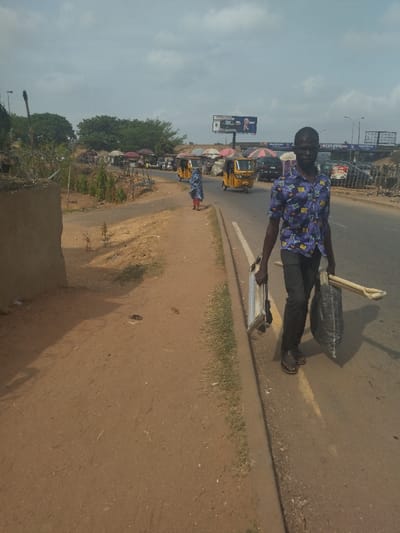 The image shows a man walking on a paved road in Maraba, Nigeria. He is wearing a blue and white patterned shirt and dark pants. He carries a metal frame and a large black bag in his right hand.  A woman wearing a blue and white dress walks on a dirt path to the left of the road. There are multiple yellow motorized three-wheeled vehicles, or tricycles, traveling on the road in the background. The road is bordered by a concrete curb. There are shops to the right side of the road. The image was taken on a partly cloudy day.  The dirt pathway on the left is partially covered with sparse vegetation. There is a concrete wall on the left, as well.  A billboard can be seen above the shops in the background.