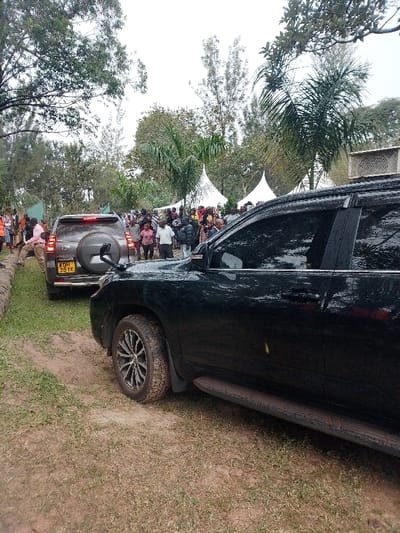 The scene is set in Marenyo-Shianda ward, Kenya, outdoors under an overcast sky. Two SUVs are positioned on a dirt path. In the immediate foreground, the right side of a dark-colored SUV, likely black, is visible, featuring a running board and multi-spoke alloy wheels with visibly muddy tires. Its windows are tinted. Behind this vehicle, a grey SUV with a rear-mounted spare tire is parked, displaying the Kenyan license plate "KDH 251K."
A significant gathering of people, estimated to be several dozens, occupies the midground and background, congregated around the path and further into a grassy area. Individuals are dressed in various casual attire. One male subject in a light pink shirt is visible near the rear of the grey SUV, appearing to interact with other individuals.
In the background, two white, conical event tents (marquees) are erected amidst a landscape dominated by diverse trees, including tall slender trees and broader palm-like trees. The ground consists of a dirt path transitioning into grassy areas. A low, stone or concrete border runs along the left edge of the dirt path.