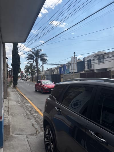The image shows a street in San Juan Cuautlancingo, Mexico, taken from a sidewalk with a building on the left side. Overhead, power lines stretch across a partly cloudy sky. A tall, narrow tree and a “VENTAS” sign are visible in the foreground. Further down the street, palm trees line the road. A red car is driving on the road, and a larger black car is parked on the right side of the frame, partially obscuring the view. Buildings line the street on both sides.