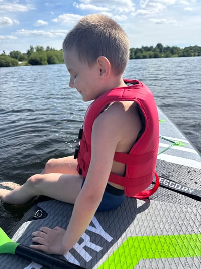 A young boy with short, light brown hair is sitting on a paddleboard in a body of water. He is wearing a red life vest over a blue bathing suit. His eyes are closed and he appears to be smiling. The paddleboard has a gray, textured surface with white and green accents. A paddle with a green blade is visible in the lower left corner of the image. The background shows a calm body of water with trees and a blue sky with white clouds. The location is the city of Ozerki, Russia.