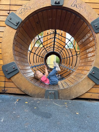 In this image, a young child is playing inside a large, circular wooden structure that appears to be part of a playground. The child, wearing a pink shirt and blue jeans, is lying on their back with their legs extended upwards, reaching out with one hand. The interior of the wooden structure is lined with horizontal wooden planks, and a black rope net forms a spherical cage within it. The exterior of the structure is made of wider wooden planks.
The wood is marked with various inscriptions, some appearing to be names and dates, with several legible as "Артем", "ЕГОР + НИКИТА БРА-", "КАТЯ ЗБИ ГИ", and "НИКИТА+". Some of these inscriptions are in black marker, while others seem to be scratched into the wood. The structure is set on a dark grey, textured surface, likely a safety matting common in playgrounds. The scene is outdoors, with glimpses of trees and a yellow structure visible through the openings in the wooden structure. The lighting suggests it is daytime. The overall impression is one of playful activity in an outdoor recreational space.