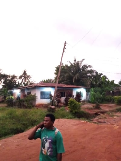 The image captures an outdoor scene in Uselu, Nigeria, under an overcast sky. In the foreground, an adult male with dark skin and short dark hair is observed from the chest up, positioned slightly to the left of the frame's center. He wears a green short-sleeved t-shirt featuring a multi-colored graphic design and a light-colored strap over his right shoulder. He is actively engaged in a phone call, holding a dark mobile phone to his right ear. The ground beneath him is an unpaved, reddish-brown dirt surface.
In the mid-ground, a single-story building with white or light-colored walls and a reddish-brown corrugated metal roof is visible. The building features several windows fitted with dark blue louvered shutters. Multiple exterior lights on the building's facade are illuminated, casting a bluish glow. A tall, slender wooden utility pole, leaning perceptibly to the right, stands in front of the building, with overhead power lines extending across the upper portion of the frame. Dense green vegetation, including trees and shrubs, surrounds and extends behind the building, interspersed with patches of grass and weeds on the unpaved ground.