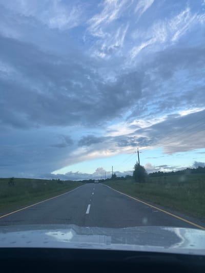 This media file depicts a road stretching into the distance, bordered by a grassy field on the left and a tree line on the right. The road has a yellow line marking its edge and white lines indicating lanes. A power line runs parallel to the road, supported by utility poles. The sky is filled with dramatic clouds, ranging from light wispy formations to darker, heavier masses, suggesting changing weather. The lighting indicates it is likely late afternoon or early evening, with the sun perhaps breaking through the clouds, casting a soft glow. The scene appears to be set in a rural or semi-rural area, given the open fields and absence of significant structures. The image is taken from inside a vehicle, as evidenced by the reflection on the right side of the frame, which shows a blurry, light-colored surface, possibly a car window or dashboard with water droplets. The overall mood is contemplative and serene, with the vast sky and open road evoking a sense of journey or travel.