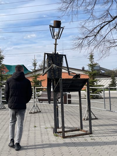 A man stands facing a metal sculpture in Urussu, Russia. The sculpture appears to be an abstract representation of a person or figure, constructed from metal rods and plates. The sculpture features a head-like structure with a cylindrical object at the top. It is standing on a paved surface with a patterned texture. Behind the sculpture are benches and some evergreen trees, with a building visible in the background. Overhead, there are tree branches and power lines against a partly cloudy sky. The man is wearing a dark jacket and gray pants and is looking at the sculpture.