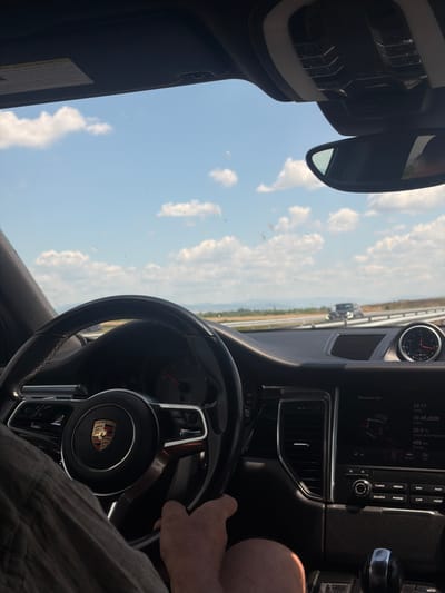 The image shows the interior of a Porsche, likely a Macan, from the perspective of the driver. The driver's hands are on the steering wheel, and part of their arm and leg are visible. The dashboard features a Porsche emblem on the steering wheel and a digital display showing trip information, time, date, outside temperature (29.0°C), and range (465 km). A clock on the dashboard shows 12:17. The exterior view through the windshield reveals a highway with other vehicles in the distance and a bright, cloudy sky. The location is identified as Загорци, Bulgaria.