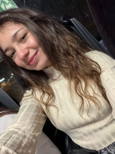 A woman poses for a selfie indoors, likely in a car, wearing a light beige ribbed top. She has long, wavy brown hair. A seatbelt is visible across her shoulder. The background shows a blurred view of a city at night through a window. She has a warm smile and appears to be in a relaxed, comfortable setting. The image has an intimate, personal feel.