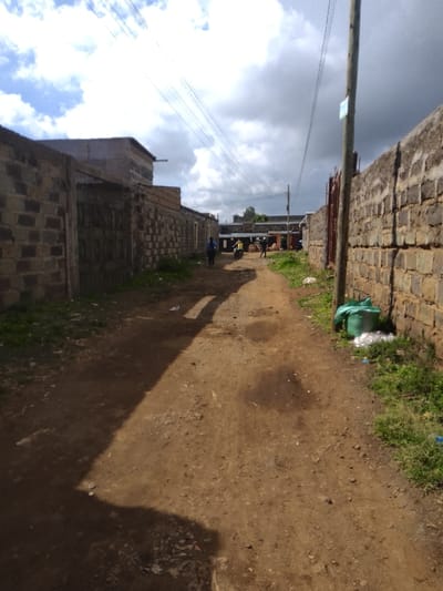 This image captures an unpaved dirt alleyway or path in Njoro ward, Kenya. The path is bordered on both sides by walls constructed of grey and brown concrete blocks.
On the left, the block wall extends into the background, partially obscuring residential or commercial structures behind it. A large, dark-colored gate or door is visible within the left wall segment further down the path. One individual, dressed in a blue top, walks away from the camera along the left side of the path.
On the right, a similar concrete block wall runs parallel to the path. A vertical wooden utility pole stands in the right foreground, supporting multiple overhead wires that traverse the sky. At the base of the right wall, a green woven sack and several scattered white plastic items are present.
Further into the background, multiple indistinct figures are visible. One individual appears to be near a light-colored two-wheeled vehicle, possibly a motorcycle or scooter, with another person seated on it. The sky above is partly cloudy, with areas of bright white clouds and darker, heavier cloud formations. Sunlight casts elongated shadows across the reddish-brown dirt path, which shows uneven terrain, depressions, and scattered small debris.