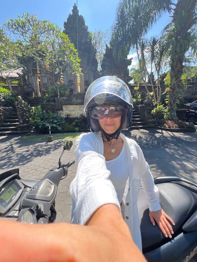 A woman wearing a helmet and white clothing is seated on a gray Yamaha scooter, possibly taking a selfie. She has her hand on the scooter seat. Behind her is a temple structure with trees and foliage, set against a blue sky. The location is Kedewatan, Indonesia, as indicated by the architectural style and vegetation. The scooter is parked on a paved surface.