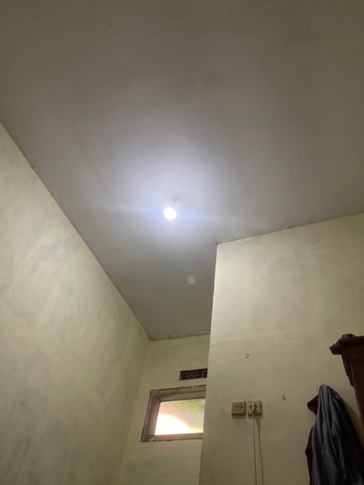 This is a low-angle, indoor photograph taken in a room in Anggaswangi, Indonesia. The ceiling is white and appears to be textured, with a single, bright lightbulb illuminating the space. The walls are a pale yellow and show signs of wear and tear, with some areas appearing rough or uneven. In the foreground, on the right side of the image, a dark wooden cabinet or wardrobe is partially visible. A dark blue jacket or piece of clothing is hanging on the wall to the left of the cabinet. On the wall, a light switch and electrical outlet are mounted. In the mid-ground, a small, rectangular window with a wooden frame is visible. The window shows a bright, indistinct light source from outside, possibly sunlight. The overall impression is of a simple, utilitarian interior space.