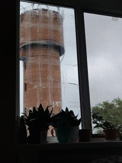The image shows a view through a window of a water tower in Okhtyrka, Ukraine. The window pane is covered with clear plastic sheeting, taped in place, distorting the view of the brick water tower. In the foreground, on a windowsill, are several potted plants, a green bottle, and a white cylindrical object. Through the right pane of the window, the tops of trees are visible against an overcast sky. The overall scene suggests a possibly damaged or improvised window covering, with everyday items on the windowsill.