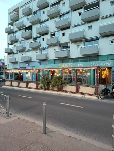 The image depicts a street-level view of a building in Canet-en-Roussillon, France. The building is a multi-story apartment complex with numerous balconies. At the street level, there is a covered outdoor seating area for a restaurant and bar named "Sanils BAR CAVE A VINS". The area is lit by string lights. Inside the establishment, patrons can be seen sitting at tables. A bicycle is parked near the entrance. The street in front of the establishment appears to be a road with a sidewalk. The overall scene suggests a commercial and residential area in a coastal town.