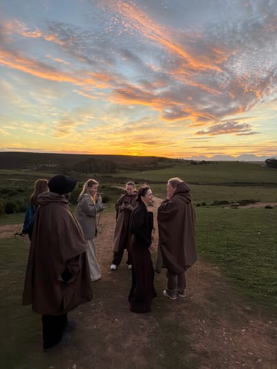 The photograph shows a group of six people standing on a grassy hilltop at sunset. They are silhouetted against a sky filled with orange and white clouds. The landscape slopes down in the distance, and the city of George, South Africa, cannot be determined from this image. All people are wearing hooded cloaks. The cloaks are brown. One person is wearing a black hat. One person is holding a phone to their face.