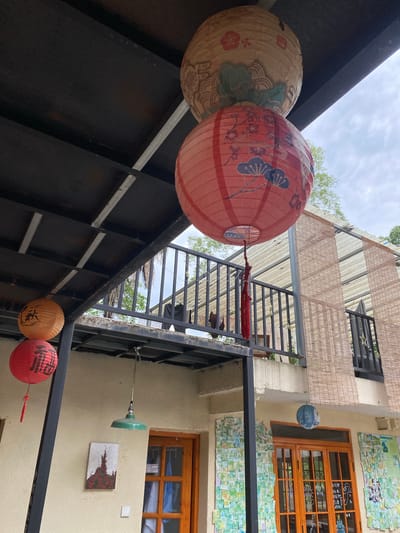This image depicts an outdoor courtyard area with several hanging lanterns and an overcast sky. In the foreground, two red lanterns are visible, one with Chinese characters that appear to be "福" (fú, meaning fortune or blessing), and another red lantern with delicate floral patterns. Above these, an orange lantern with Chinese characters that appear to be "秋" (qiū, meaning autumn) is hanging. Further up, two more lanterns are stacked, one being a red lantern adorned with blue floral designs and a red tassel, and the other being a larger, beige lantern with intricate floral patterns. The lanterns are suspended from a dark, sloped overhang.

The background reveals a two-story building with a balcony railing. Bamboo blinds are hung over windows on the upper floor. A green industrial-style pendant lamp hangs from the overhang, positioned above a framed artwork on the wall. The wall is a light beige color and is decorated with numerous small, colorful papers, possibly photographs or postcards. Two wooden framed glass doors are visible on the ground floor. The overall impression is of a charming, possibly cultural or artistic, space. The image is consistent with an outdoor dining or gathering area in a scenic location. Given the context of Dujiangyan City, China, this scene might be from a traditional guesthouse, teahouse, or a shop catering to tourists.