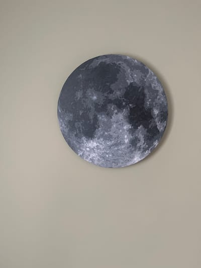 A circular painting of the moon hangs on a light beige wall. The painting is rendered in various shades of gray and white, depicting the moon's textured surface. The painting is positioned in the upper right quadrant of the image. The wall appears to be a matte finish. No other objects or people are visible in the frame. The location is Staryzhivotinnoye, Russia.