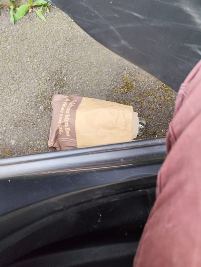 The image shows a brown paper bag lying on a gray asphalt surface, partially visible under the open rear door of a car. The bag is partially obscured by the car's door and a burgundy garment. Text on the bag reads "C'est un bien fou de voler son sac". The coordinates 45.782, 3.124 locate the scene in the city of Clermont-Ferrand, France. The location is most likely near a street or parking lot. The scene suggests a discarded bag, perhaps left behind by a passerby.
