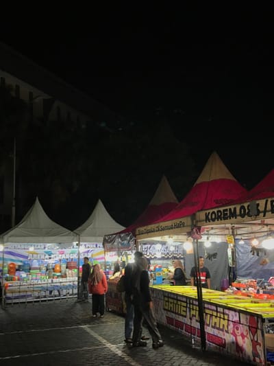The image shows a night market in the Kecamatan Batu district of Indonesia, located at coordinates -7.867 latitude and 112.514 longitude. The market is set up under several large white and red tents with various stalls displaying brightly colored goods. There are several people walking through the market, browsing the goods. The tents have banners in Indonesian and English. One banner reads, "Bisa Dipakai Di Semua Hape" which translates to "Can be used on all phones." Other banners are visible, but their text is not readable. The market appears to be well-lit, and the environment is bustling with activity.