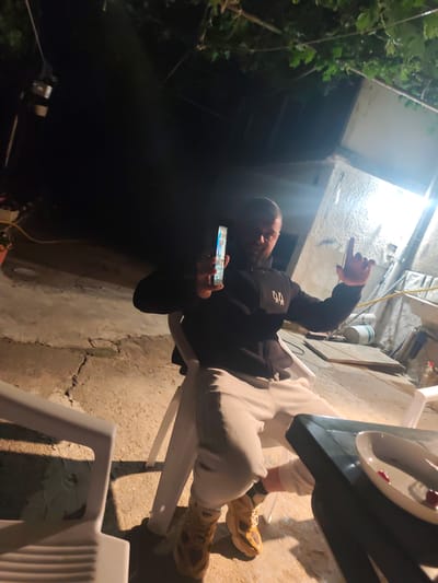 A man sits in a white plastic chair outside at night. He holds a lit smartphone in his left hand and points with his right index finger. The man wears a black zip-up jacket, light-colored pants, and tan sneakers. To his right, a dark-colored table holds a white plate with remnants of food. The background shows a concrete patio area with cracks and a white building illuminated by a bright light. Green vines hang overhead. The location is in Balgarchevo, Bulgaria.