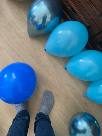 The image shows a pair of legs wearing grey socks and blue jeans, standing on a wooden floor. The legs are positioned between a large, bright blue balloon on the left and a cluster of lighter blue and metallic blue balloons on the right. One metallic blue balloon is partially visible in the bottom right corner. The balloons are scattered across the floor. In the background, a dark wooden cabinet with a metal drawer pull is visible. The scene appears to be indoors, likely in a residential setting, given the wooden floor and furniture. There is no specific indication of the location being Osek, Czechia.