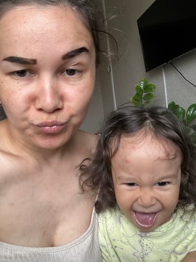 A selfie shows a woman and a young child. The woman has dark hair pulled back and is looking at the camera. The child is positioned above her, also facing the camera, with their tongue sticking out. The location appears to be indoors, possibly a residential setting, based on the presence of a wall and a plant in the background. There's no information to definitively place the scene in the city of Uray, Russia.