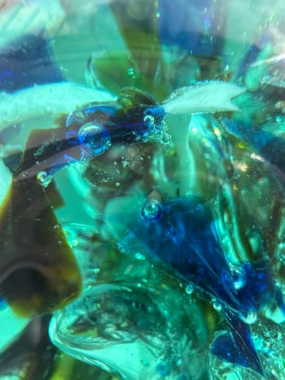 The image displays an abstract, close-up view of colorful glass shards and bubbles, predominantly in shades of blue and green. The glass appears to be fractured and suspended in a translucent medium, possibly resin or liquid. The lighting creates highlights and refractions, emphasizing the textures and depth of the material. There are no discernible people, actions, or interactions depicted. The scene is an inanimate composition of glass fragments, lacking any context related to a specific city or location like Barvikha, Russia.
