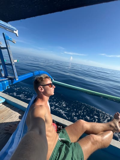 A man is lying on a boat in Seririt, Indonesia. He is shirtless, wearing green shorts and sunglasses, and has a light blue and white striped towel beneath him. His legs are crossed and propped up on the side of the boat. The boat has blue and green railings. The sea is visible in the background with a clear blue sky above.