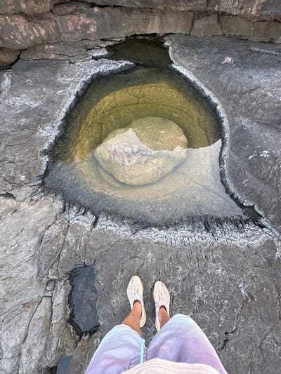 The image shows a person's feet standing on dark, wet rocks near a tidal pool. The tidal pool is filled with clear, yellowish water and contains a large, smooth rock at its center. The surrounding rocks are rough and appear to be volcanic or sedimentary in origin, with visible cracks and textured surfaces. The area is outdoors, with the lighting suggesting it is daytime. Based on the description of the location, this scene is likely situated in the coastal region of Lastva Grbaljska, Montenegro, a popular area known for its rocky shores and natural formations.