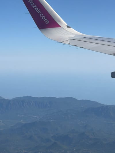 An aerial view shows a portion of an airplane wing with the text "wizzair.com" printed on a purple section. The wing is angled upwards and to the right, with the clear blue sky filling the background. Below the wing, a hazy, mountainous landscape is visible. The mountains appear layered and are covered in dark green vegetation, suggesting a natural, undeveloped area. The image's perspective indicates the photograph was taken from inside the airplane. The location of Donja Cijevna, Montenegro, is not visually identifiable from the provided image.