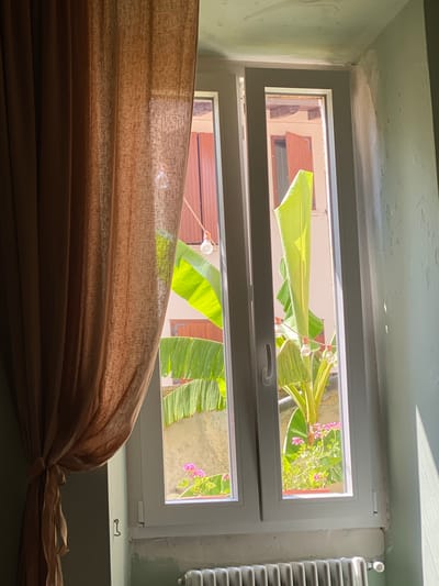 The image shows a window with a light brown curtain partially drawn to the left. Through the window, a banana tree with large green leaves and small pink flowers in a terracotta pot are visible. In the background, there is a building with wooden shutters and some outdoor lights. The scene is lit by sunlight casting shadows. The window frame is white, and the wall around it is painted in a muted green color. A white radiator is visible below the window. The image appears to be taken from inside a room, possibly in Lonato del Garda, Italy, given the architectural style and vegetation.